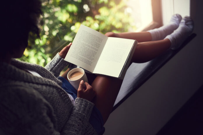 Shot of an unidentifiable young woman reading a book while enjoying a cup of coffee at home