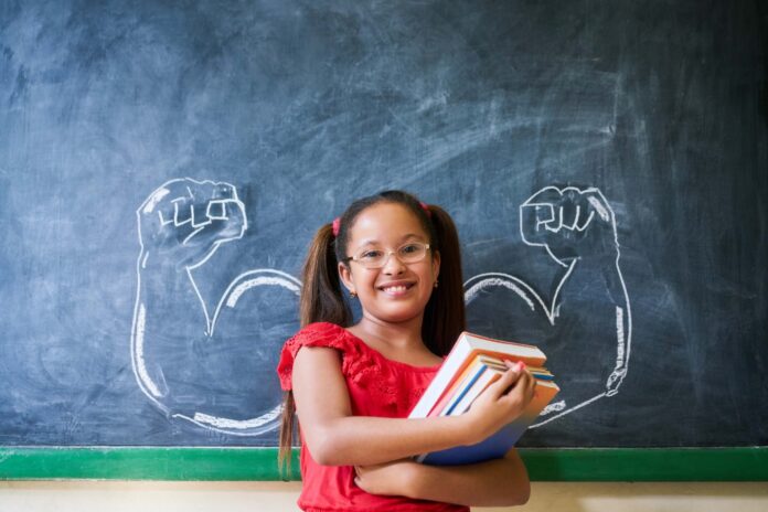 Concept on blackboard at school. Intelligent and successful hispanic girl in class. Portrait of female child smiling, looking at camera, holding books against drawing of muscles on blackboard (Concept on blackboard at school. Intel